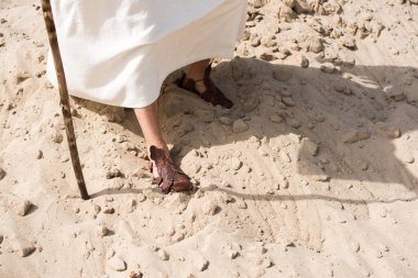 cropped image of Jesus in robe and sandals walking on sand with wooden staff in desert