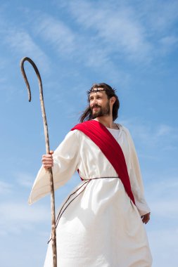 cheerful Jesus in robe, red sash and crown of thorns standing with wooden staff in desert clipart