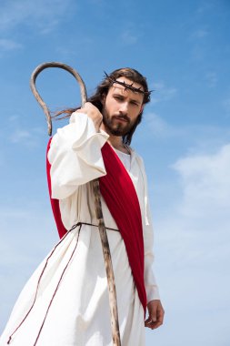 low angle view of Jesus in robe, red sash and crown of thorns standing with wooden staff in desert and looking at camera