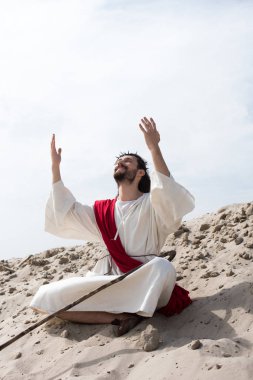 smiling Jesus in robe, red sash and crown of thorns sitting in lotus position with raised hands and talking with god on sand in desert clipart