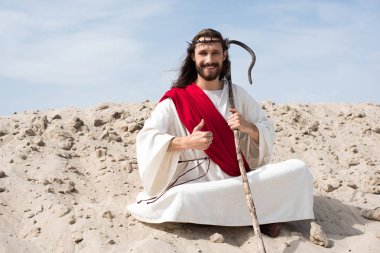 İsa'nın lotus pozisyonu kum üzerinde oturan, personel holding ve çölde başparmak gösterilmesini gülümseyen