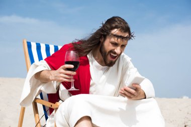 angry Jesus resting on sun lounger with glass of wine and screaming at smartphone in desert clipart
