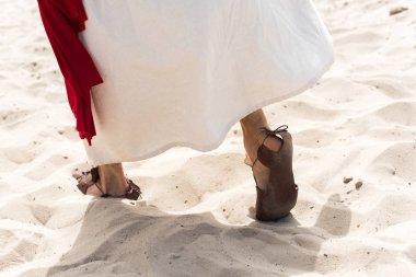 cropped image of Jesus in robe, sandals and red sash walking on sand in desert clipart