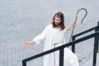 high angle view of cheerful Jesus in robe and crown of thorns walking on stairs with staff and showing hand clipart