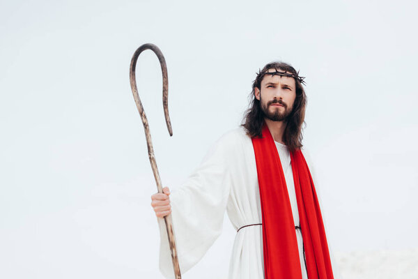 low angle view of Jesus in robe, red sash and crown of thorns standing with wooden staff in desert