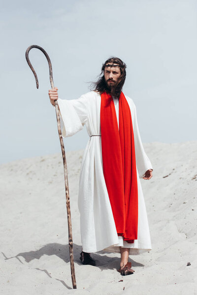 Jesus in robe, red sash and crown of thorns standing with wooden staff in desert and looking away