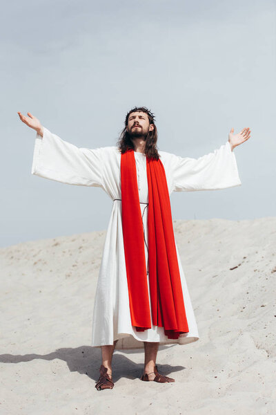 Jesus in robe, red sash and crown of thorns standing with raised hands and talking with god in desert