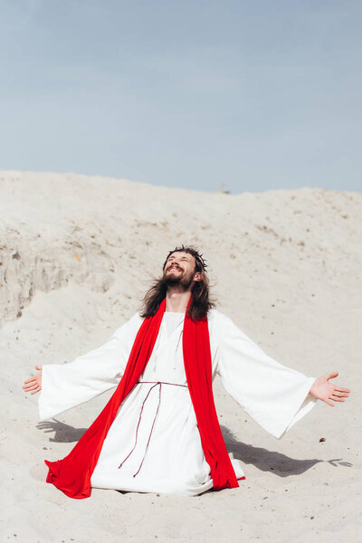 happy Jesus in robe, red sash and crown of thorns standing on knees with open arms in desert