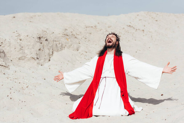laughing Jesus in robe, red sash and crown of thorns standing on knees with open arms in desert