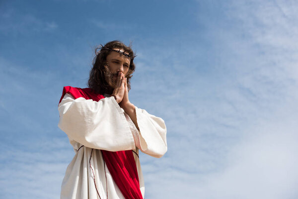 low angle view of Jesus in robe and red sash praying against blue sky