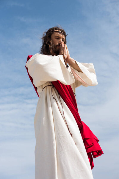 Низкий угол зрения Иисуса в халате, красная лента и терновый венец, держа четки и молясь против голубого неба
