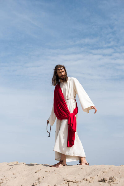 Иисус в мантии, красной ленте и терновом венце, держа чётки и идя по песчаному холму в пустыне
