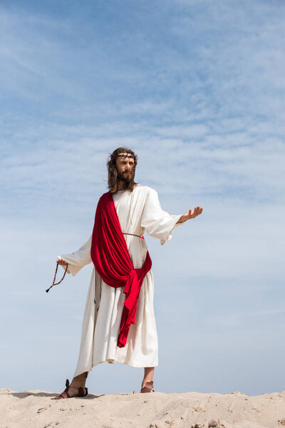 Jesus in robe, red sash and crown of thorns holding rosary and standing with open arms in desert