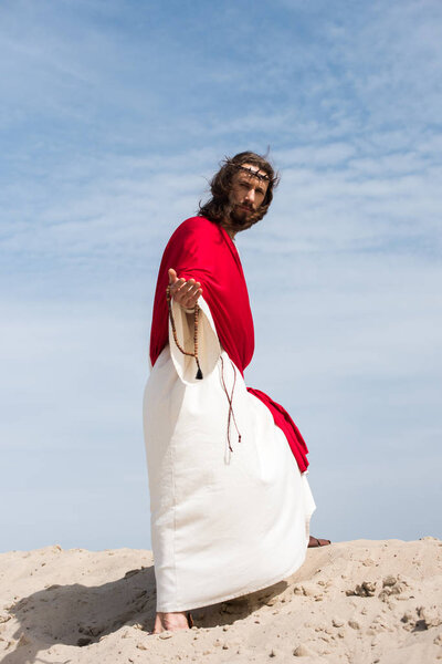 Jesus in robe, red sash and crown of thorns holding rosary and giving hand in desert