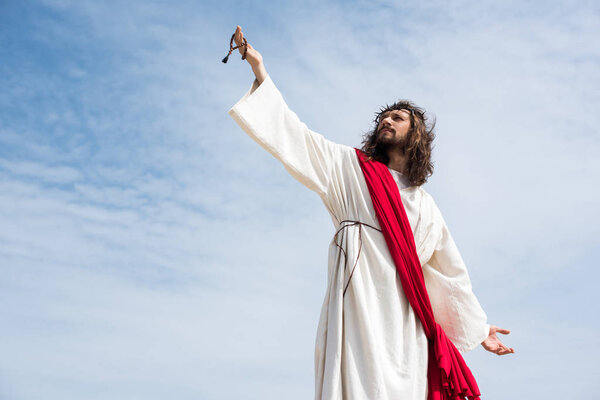 low angle view of Jesus in robe, red sash and crown of thorns holding rosary in raised hand against blue sky