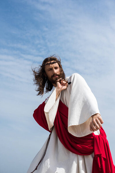 low angle view of Jesus in robe, red sash and crown of thorns having fun and dancing with rosary against blue sky