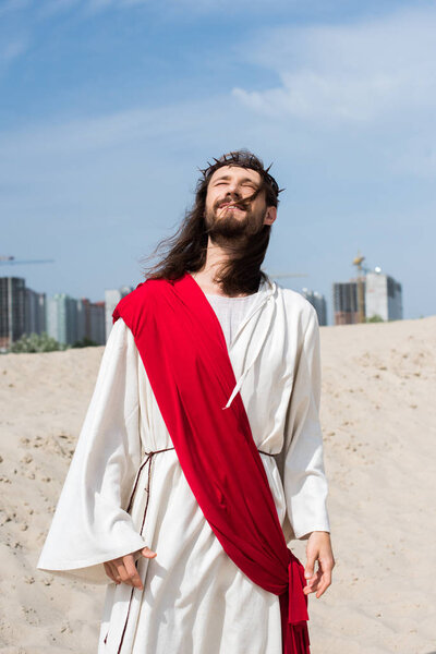 Jesus in robe, red sash and crown of thorns standing on sand with closed eyes, buildings on background