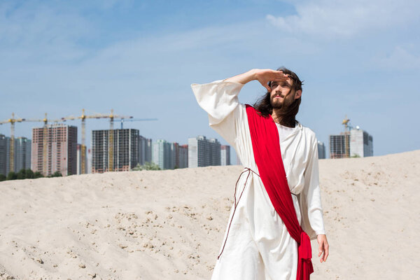 Иисус в мантии, красной ленте и терновом венце стоял на песке и смотрел на здания на заднем плане
