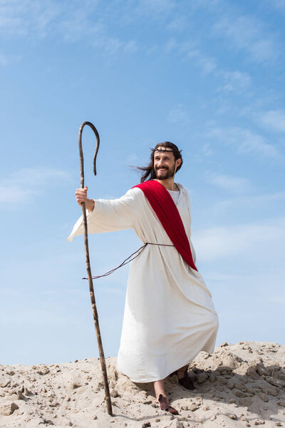 smiling Jesus in robe, red sash and crown of thorns standing with wooden staff in desert