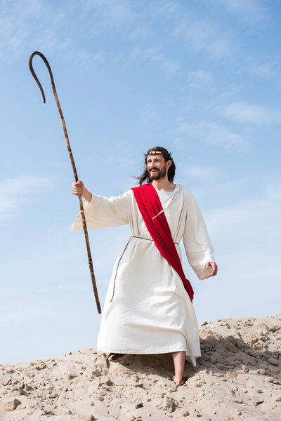 счастливый Иисус в мантии, красной ленте и терновом венце, стоящий с деревянным посохом в пустыне и смотрящий в сторону
