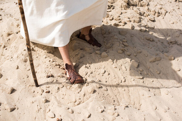 обрезанный образ Иисуса в халате и сандалиях, идущих по песку с деревянным посохом в пустыне
