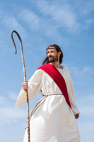 веселый Иисус в мантии, красной ленте и терновом венце, стоящем с деревянным посохом в пустыне

