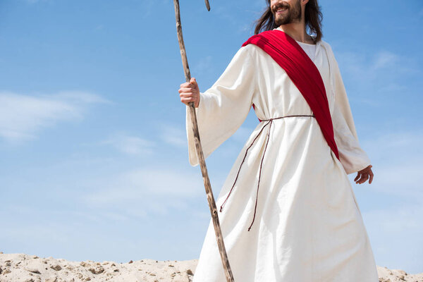 cropped image of smiling Jesus in robe and red sash standing with wooden staff in desert
