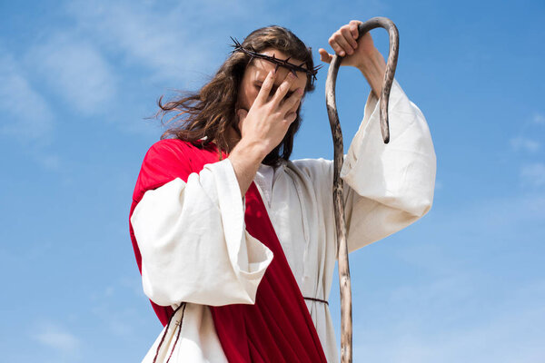 low angle view of Jesus in robe, red sash and crown of thorns standing with wooden staff in desert and touching forehead