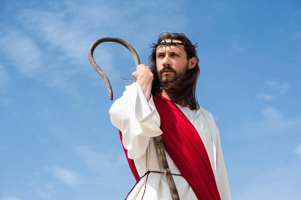 Jesus in robe, red sash and crown of thorns standing with wooden staff against blue sky and looking away