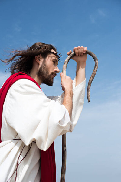 side view of Jesus in robe and crown of thorns leaning on wooden staff against blue sky