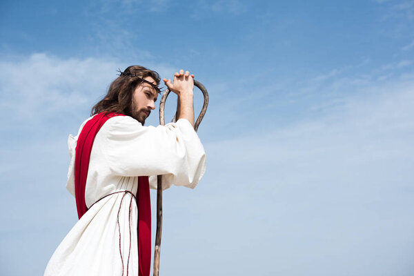 side view of Jesus in robe, red sash and crown of thorns leaning on wooden staff against blue sky