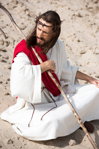 Jesus in robe, red sash and crown of thorns sitting in lotus position on sand in desert and looking away