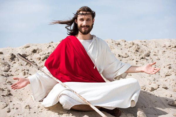 smiling Jesus in robe, red sash and crown of thorns sitting in lotus position with open arms on sand in desert