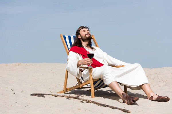 Jesus in robe and red sash resting on sun lounger with glass of red wine in desert