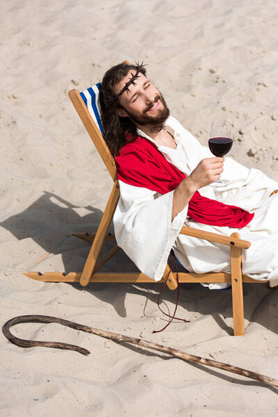 smiling Jesus in robe and red sash resting on sun lounger with glass of red wine in desert