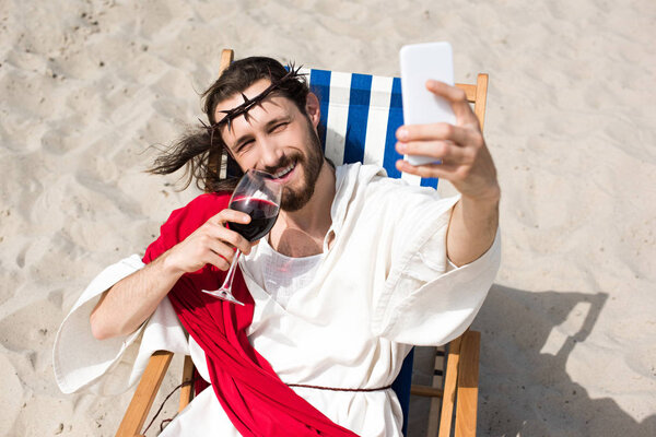 smiling Jesus resting on sun lounger, drinking wine and taking selfie with smartphone in desert