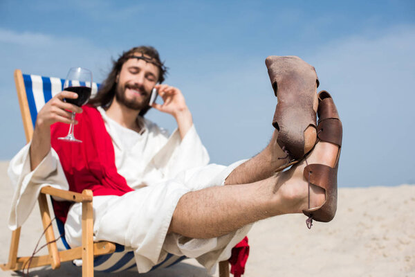 smiling Jesus resting on sun lounger with glass of wine and talking by smartphone in desert