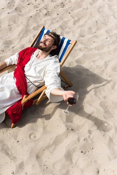 high angle view of happy Jesus in robe and red sash relaxing on sun lounger with glass of red wine in desert