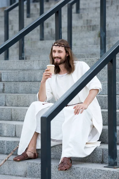 smiling Jesus in robe and crown of thorns sitting on stairs and holding disposable coffee cup on street