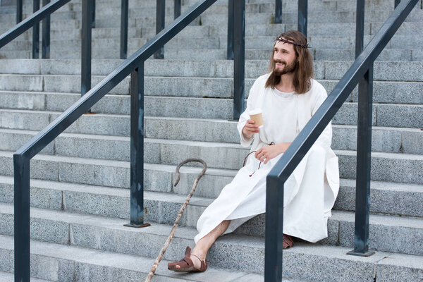 cheerful Jesus in robe and crown of thorns sitting on stairs and holding disposable coffee cup on street