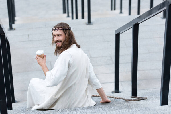 back view of smiling Jesus in robe and crown of thorns sitting on stairs and holding disposable coffee cup on street
