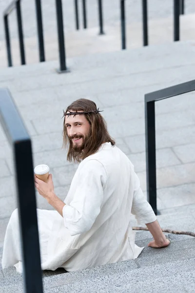 rear view of smiling Jesus in robe and crown of thorns sitting on stairs and holding disposable coffee cup on street