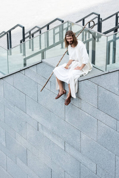 smiling Jesus in robe and crown of thorns sitting on staircase side, holding disposable coffee cup and looking down