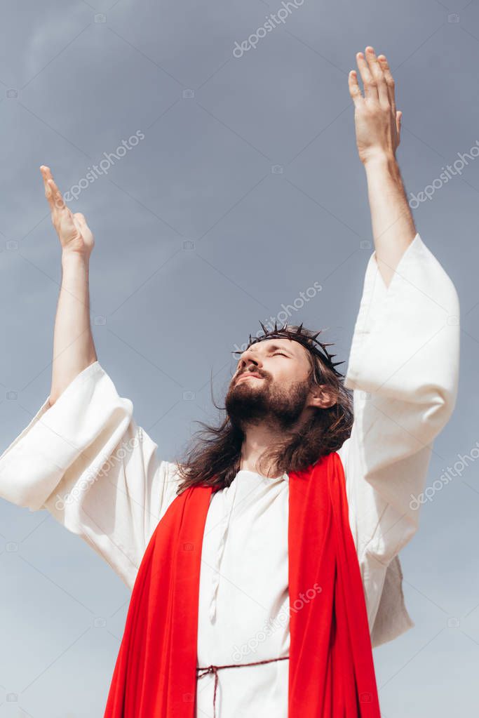 low angle view of Jesus in robe, red sash and crown of thorns standing with raised hands against grey sky