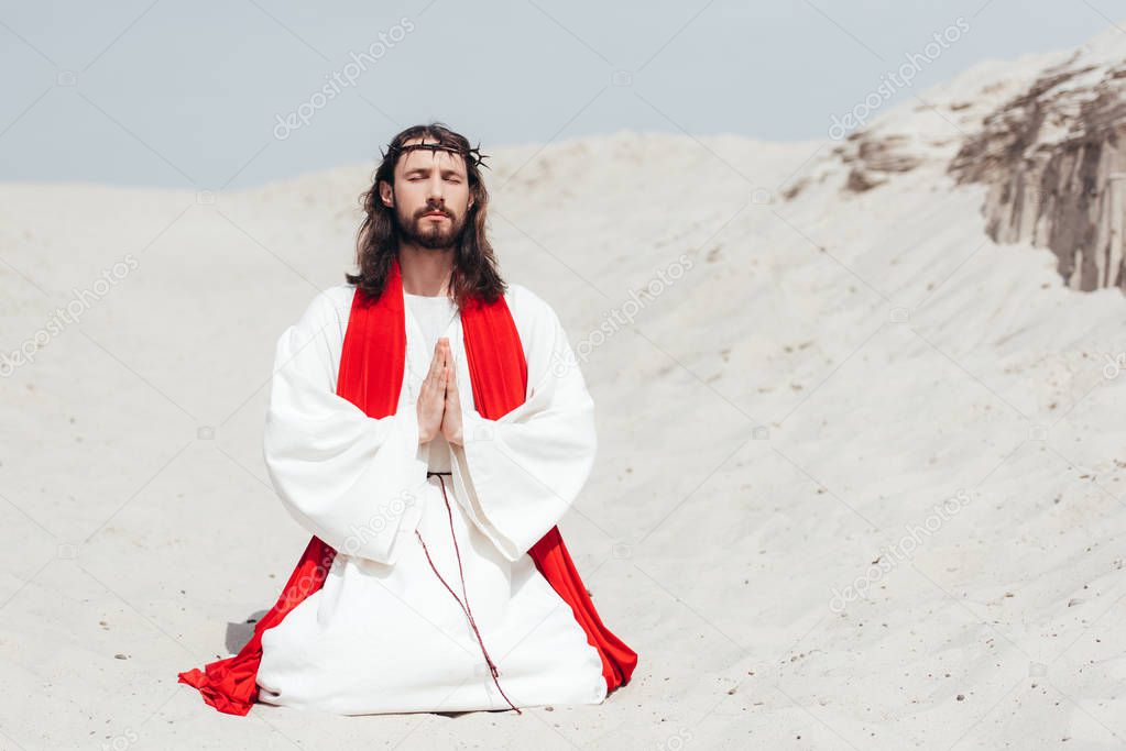 Jesus with closed eyes standing on knees on sand and praying in desert