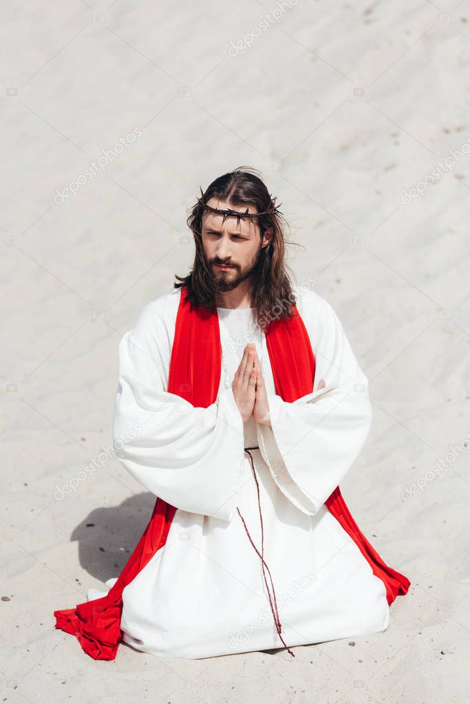 Jesus in robe, red sash and crown of thorns standing on knees and praying in desert, looking away