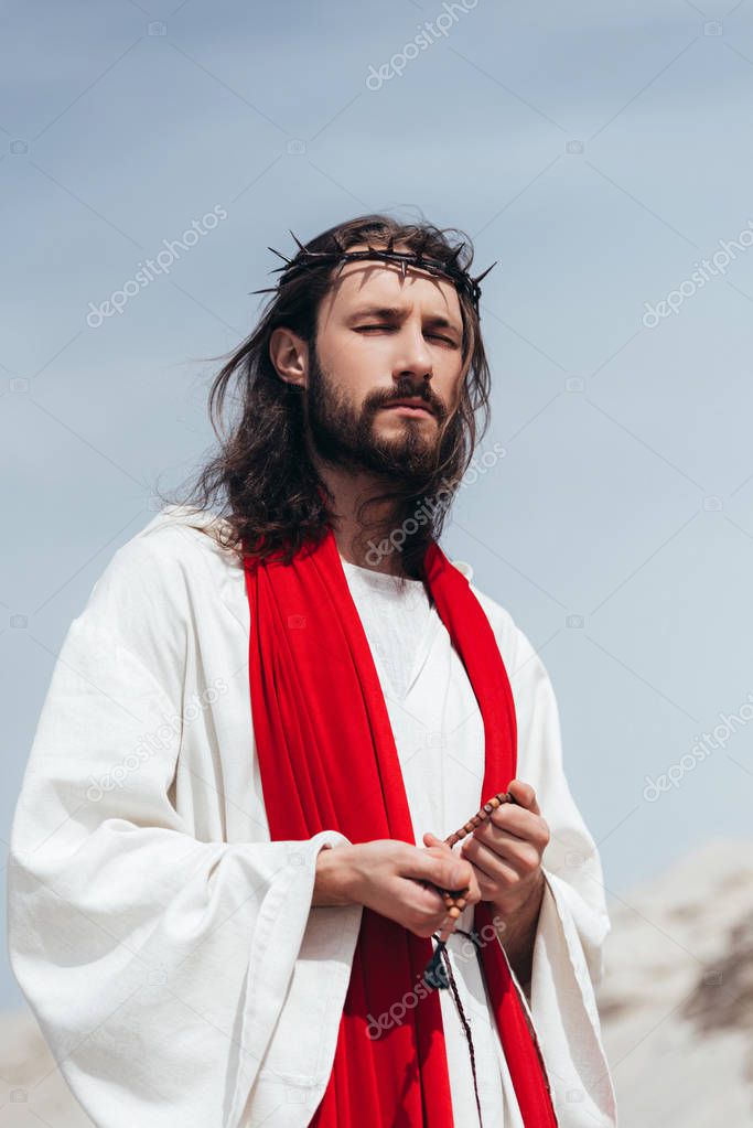 Jesus in robe, red sash and crown of thorns holding rosary and praying with closed eyes in desert