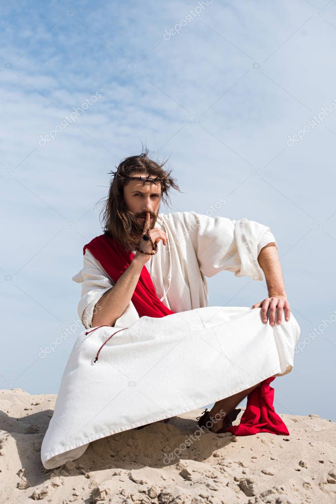 Jesus in robe, red sash and crown of thorns holding rosary and showing silence sign in desert