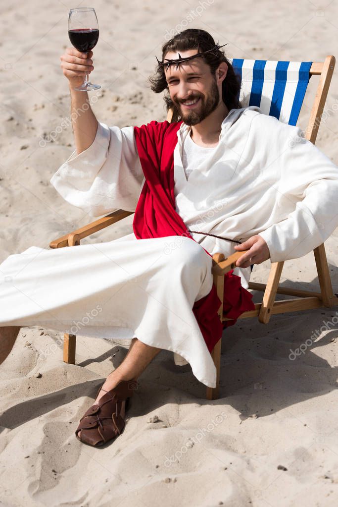 cheerful Jesus in robe and red sash resting on sun lounger and showing glass of red wine in desert