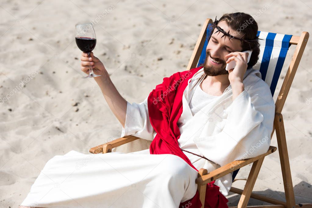 smiling Jesus resting on sun lounger with glass of wine and talking by smartphone in desert